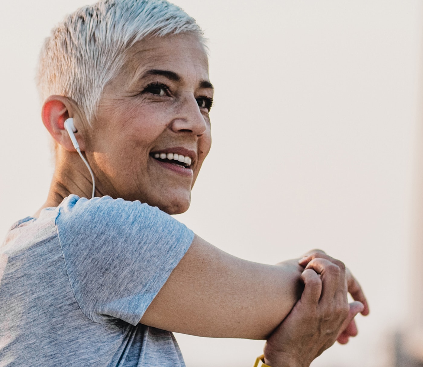 Smiling woman wearing earbuds stretching outside.