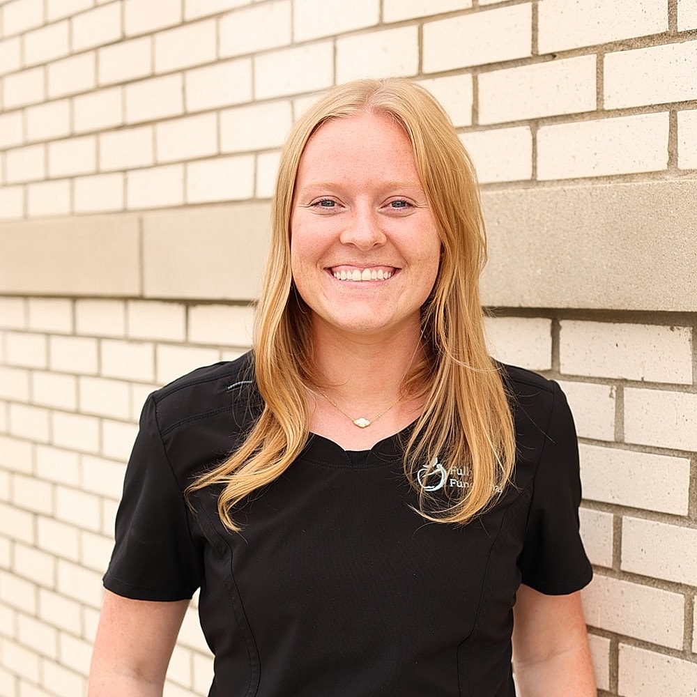 Woman in black scrubs smiling by brick wall.