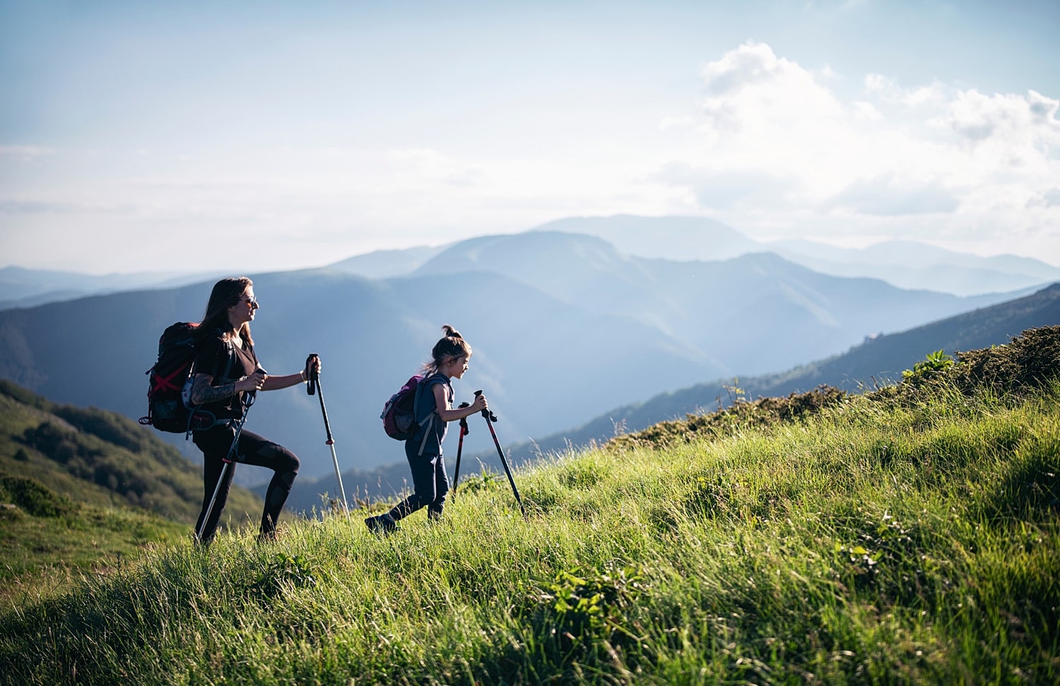 Hiking mother and daughter on grassy mountain trail.